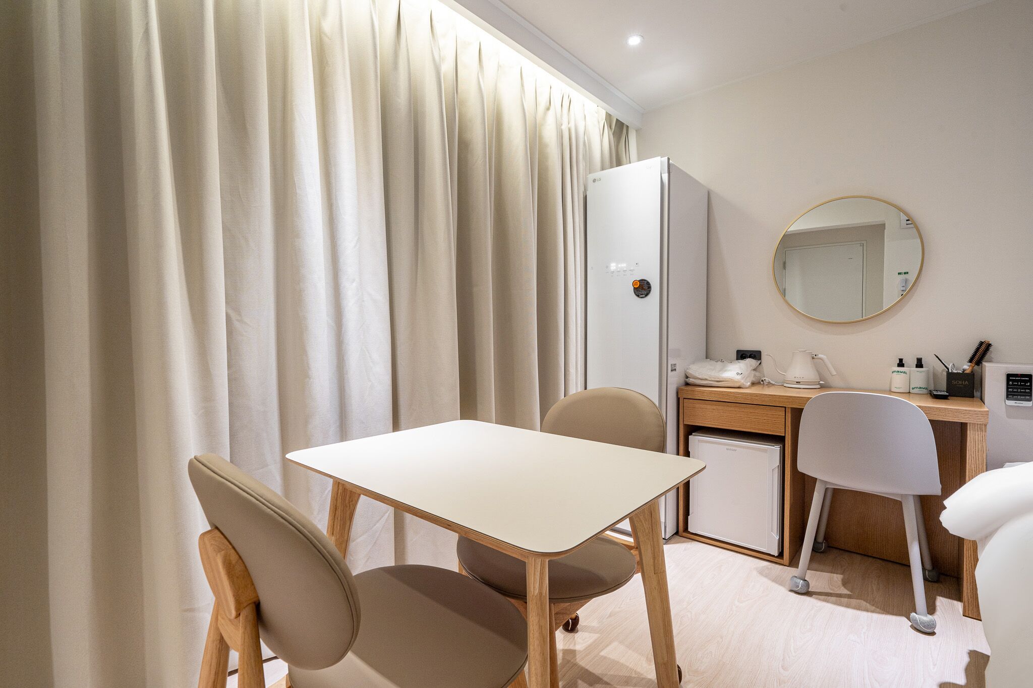 A hotel room with a dining table, two chairs, a small refrigerator, a desk with a chair, a mirror, and a curtain.