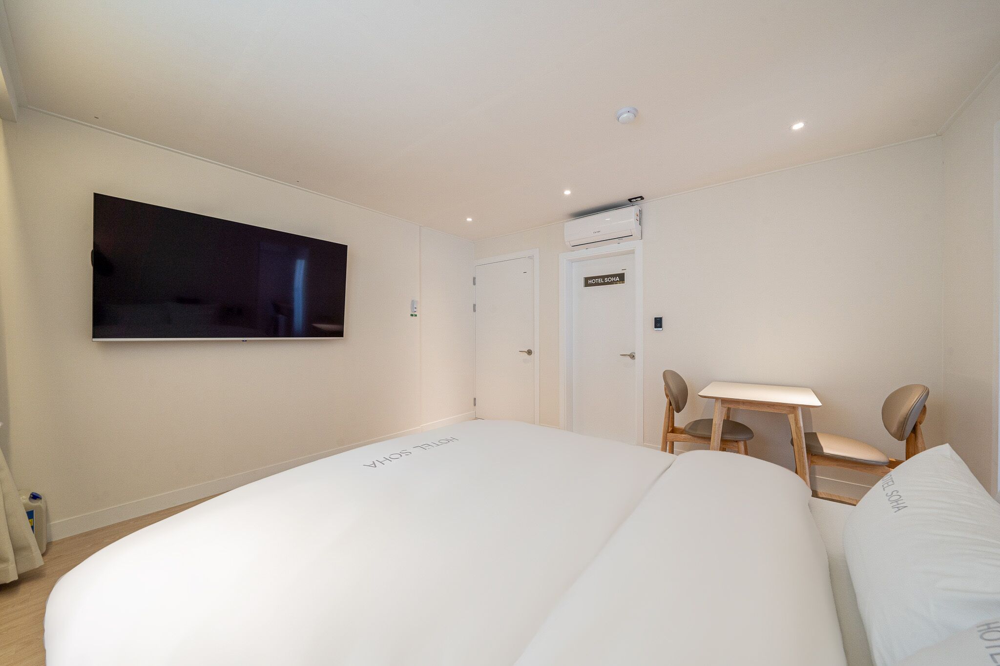 Ein Hotelzimmer mit einem großen Bett, einem an der Wand befestigten Fernseher, einem Schreibtisch mit zwei Stühlen und einer Tür mit der Aufschrift 'HOTEL RONA'.
