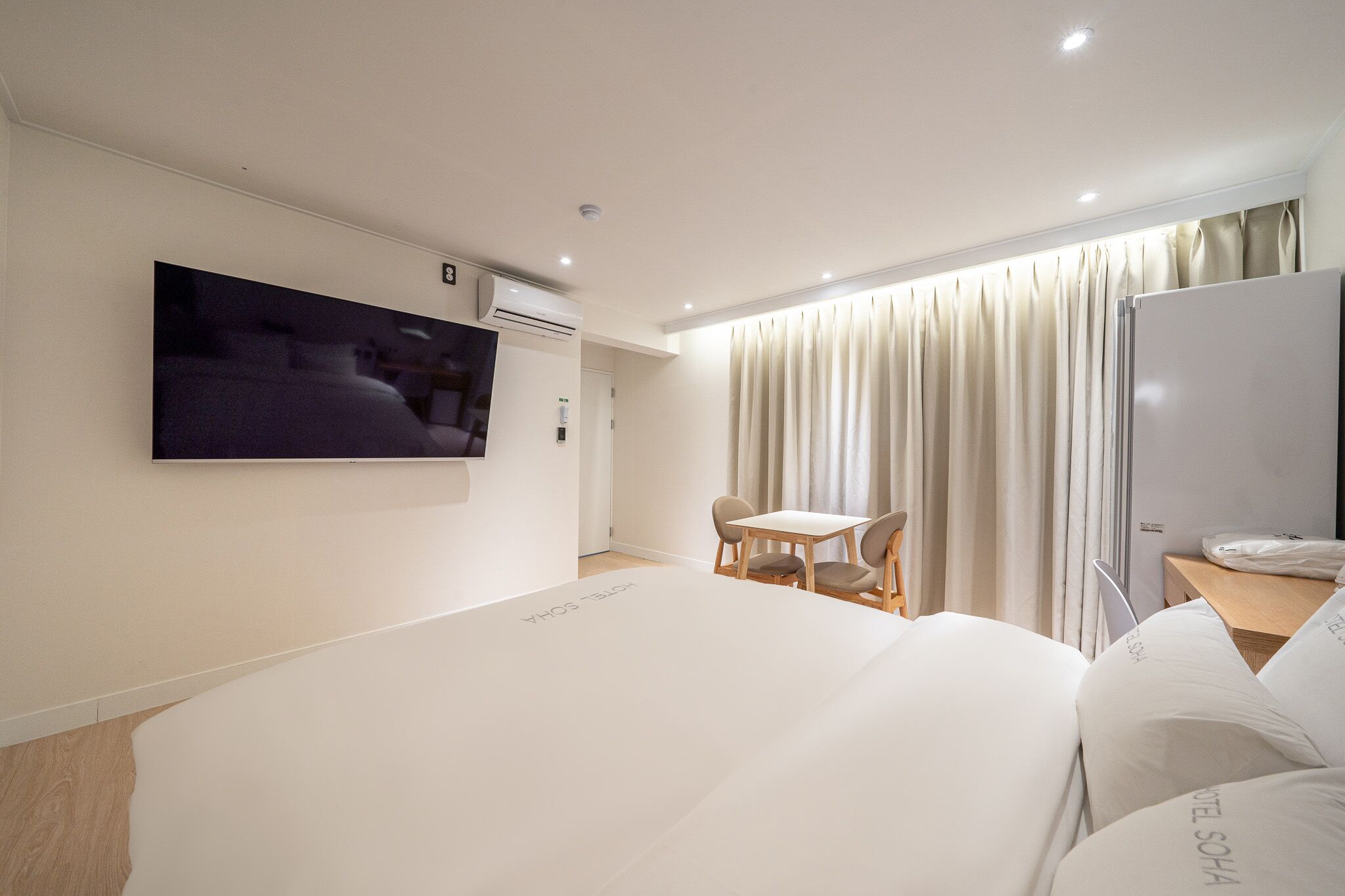 A hotel room with a large bed, a TV mounted on the wall, a small table with chairs, and a window with curtains.