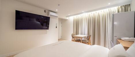 A hotel room with a large bed, a TV mounted on the wall, a small table with chairs, and a window with curtains.
