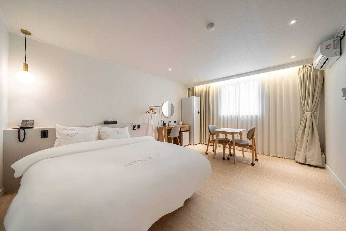 A hotel room with a large bed, a desk with a chair, a small table with a chair, a telephone, and an air conditioning unit.