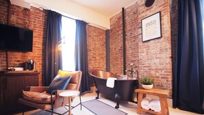 Una sala de estar moderna con pared de ladrillo, un ventanal con cortinas, un sillón de cuero, una mesa redonda para café, una bañera negra y una pequeña mesa de madera con toallas.
