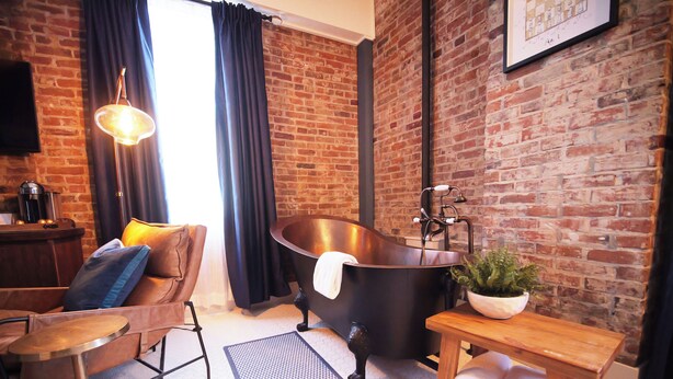 Un baño moderno con una bañera independiente grande, un banco de madera con toallas, una cafetera y una pared de ladrillo.