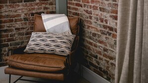 A corner room with a brick wall, a leather chair with a patterned throw, and a curtain.