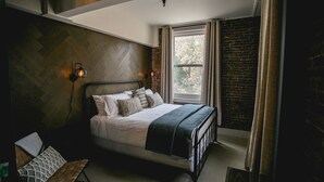 Un dormitorio con pared de patrón espiga, cama con cubrecama blanco y varios almohadones, mesita de noche con lámpara y ventana con cortinas.