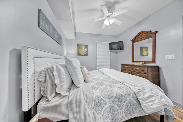Une chambre à coucher comprenant un lit, une commode, un ventilateur de plafond, un téléviseur fixé au mur et un miroir décoratif.