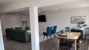 A modern living room with a grey sofa, a dining table with chairs, and a TV mounted on the wall.