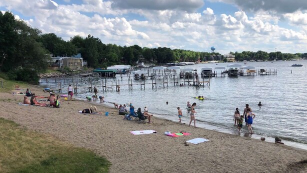 A beach with people sunbathing and swimming, a dock area with boats, and a water tower in the background.