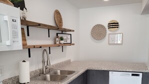 A modern kitchen with a granite countertop, stainless steel sink, and a built-in dishwasher. There are wooden shelves with decorative items and a microwave above the counter.