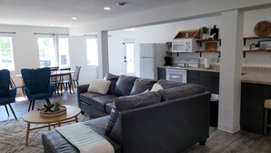 A modern living room with a grey sofa, blue armchairs, and a small coffee table. A kitchen with white appliances and a dining area with a table and chairs is visible.