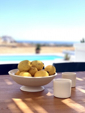 En skål med citroner står på ett träbord, och i bakgrunden syns en strand och en pool.