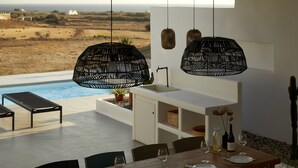 Ein Essbereich mit Blick auf den Ozean, einem Pool und einem minimalistischen Design mit schwarzen, gewebten Pendelleuchten.