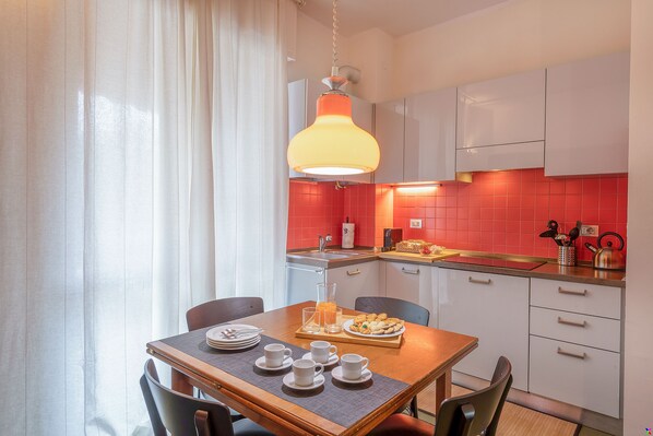 A modern kitchen with white cabinets, a red backsplash, and a dining area with a wooden table set for breakfast. - BnButler - Brivio 21 (Milan)