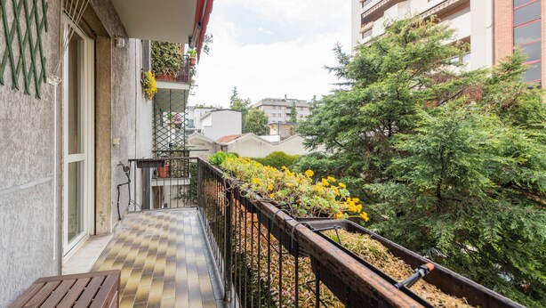 A balcony with a wooden bench, a metal railing, and a view of a garden with yellow flowers and green trees. - BnButler - Brivio 21 (Milan)