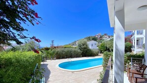 Une piscine entourée de verdure et de bâtiments blancs, avec un ciel bleu dégagé au-dessus.