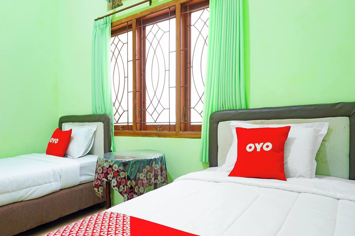 Two single beds with red pillows, a small round table with a floral tablecloth, and a window with green curtains.
