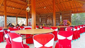 A covered outdoor dining area with round tables and red chairs with white sashes.