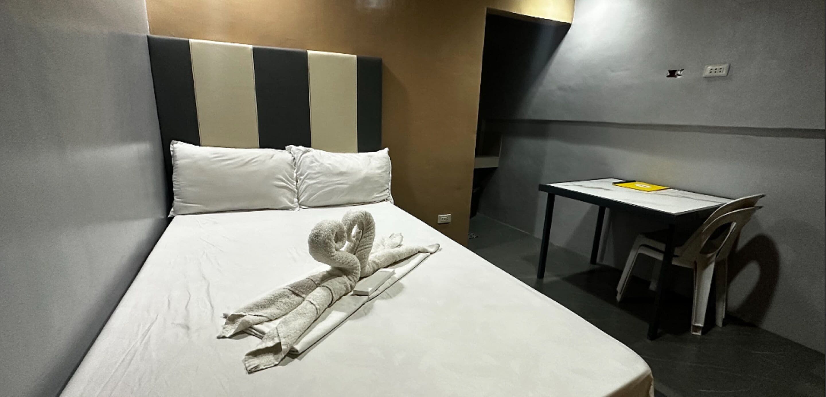 Hotelkamer met een bed, een netjes opgemaakt bed met een handdoek in de vorm van een zwaan, een klein tafeltje met een stoel, en een effen wand met een stopcontact.