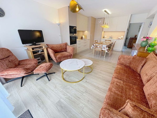 Un salon moderne comprenant un ensemble de canapés, un téléviseur, un coin repas et une cuisine en arrière-plan.