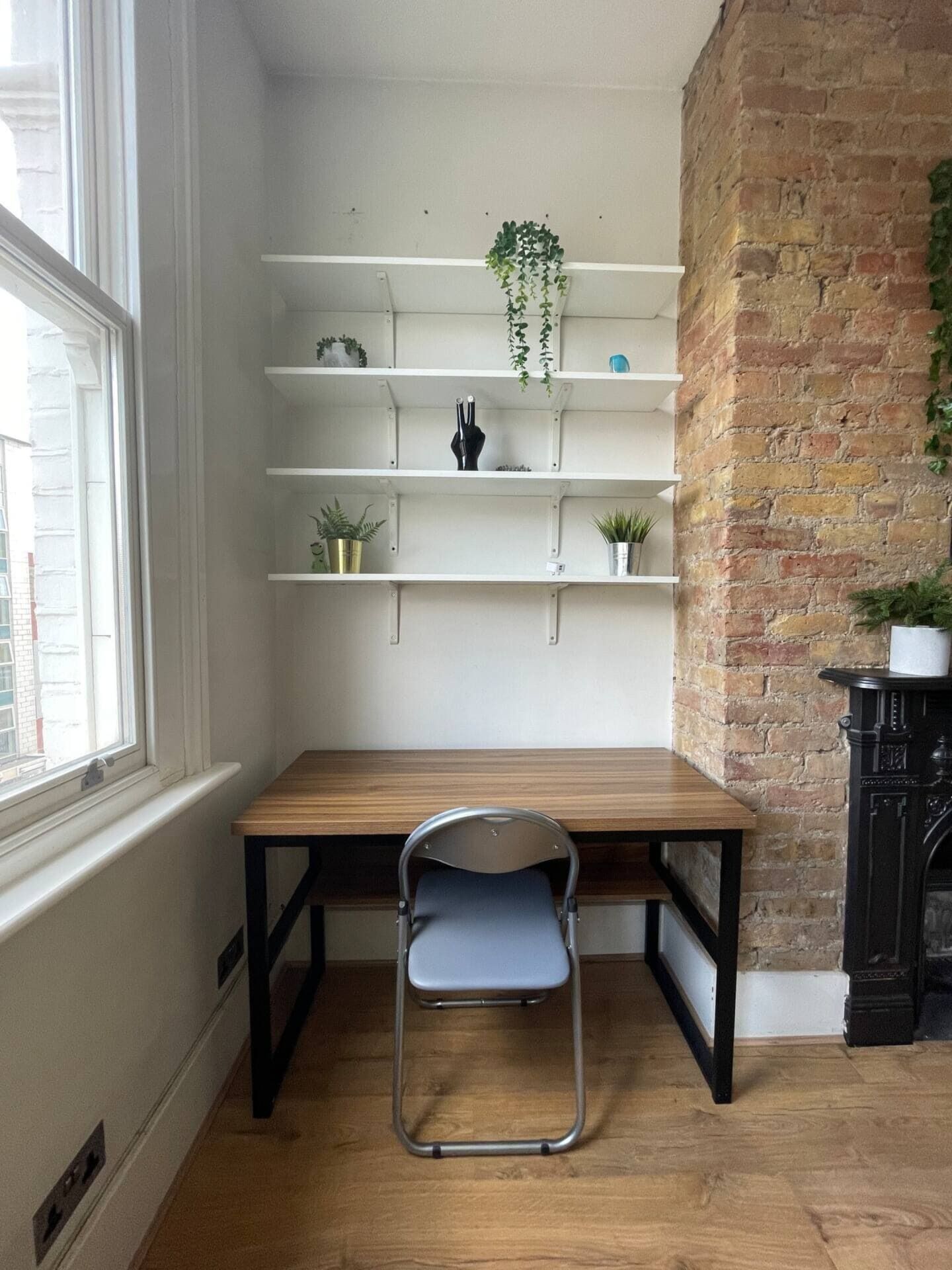 A corner room with a wooden table, a foldable chair, and white shelving units with plants.