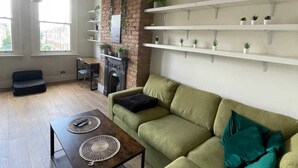 Una sala de estar moderna con un sofá verde, una mesa de centro de madera y una chimenea. Hay estantes blancos en la pared y una lámpara colgante.