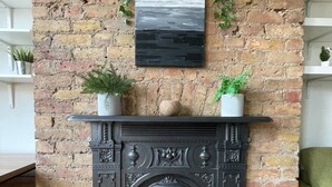 Una chimenea de ladrillo con molduras decorativas, dos plantas en macetas sobre la repisa, y una obra enmarcada arriba.
