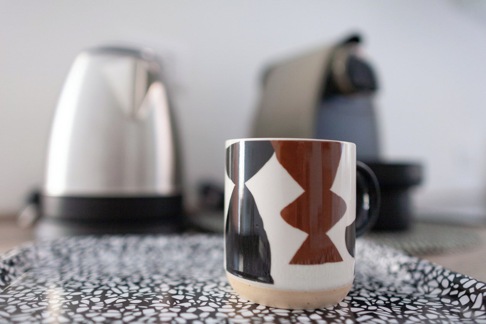 A mug with a geometric pattern on a patterned tablecloth, a kettle, and a chair in the background.