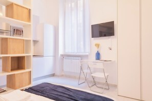 A modern, well-lit room with a white color scheme, featuring a wooden bookshelf, a desk with a chair, a radiator, and a mounted TV. - BnButler - Chieti 5 (Milan)