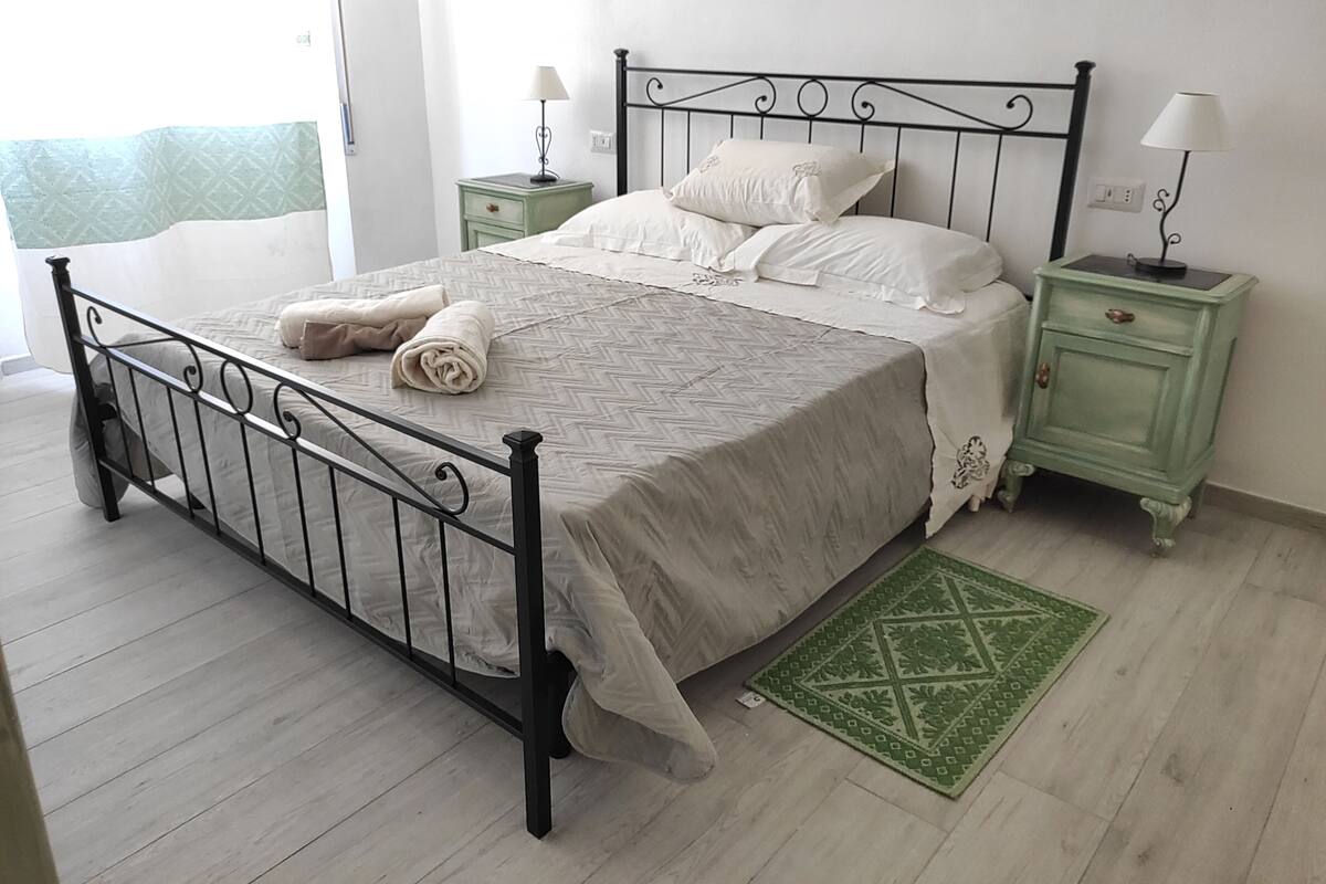 Un lit bien fait avec un cadre en métal, une table de chevet verte avec une lampe, et un tapis à motifs verts sur un plancher en bois clair.