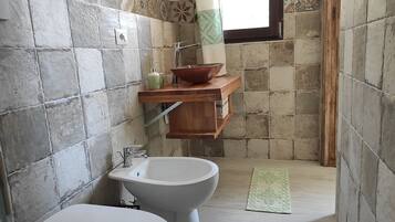 Un baño con inodoro, bidé y un estante de madera con un cuenco y botellas.