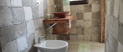 A bathroom with a toilet, bidet, and a wooden shelf with a bowl and bottles.