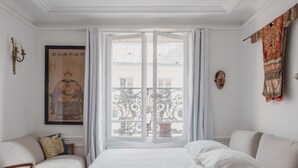 Une chambre avec une grande fenêtre, un lit, deux chaises, une lampe fixée au mur, un tableau encadré et un masque décoratif.
