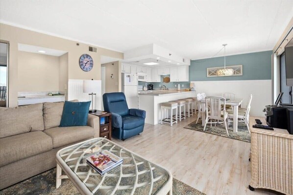 A modern living room with a sofa, armchair, and glass coffee table. A kitchen with white cabinets and a dining area with white chairs is visible in the background. - Bayfront Condo with Beautiful Views and Pool (Ocean City)