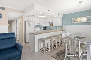 A modern kitchen with white cabinets, a central island, bar stools, and a dining table with chairs. - Bayfront Condo with Beautiful Views and Pool (Ocean City)