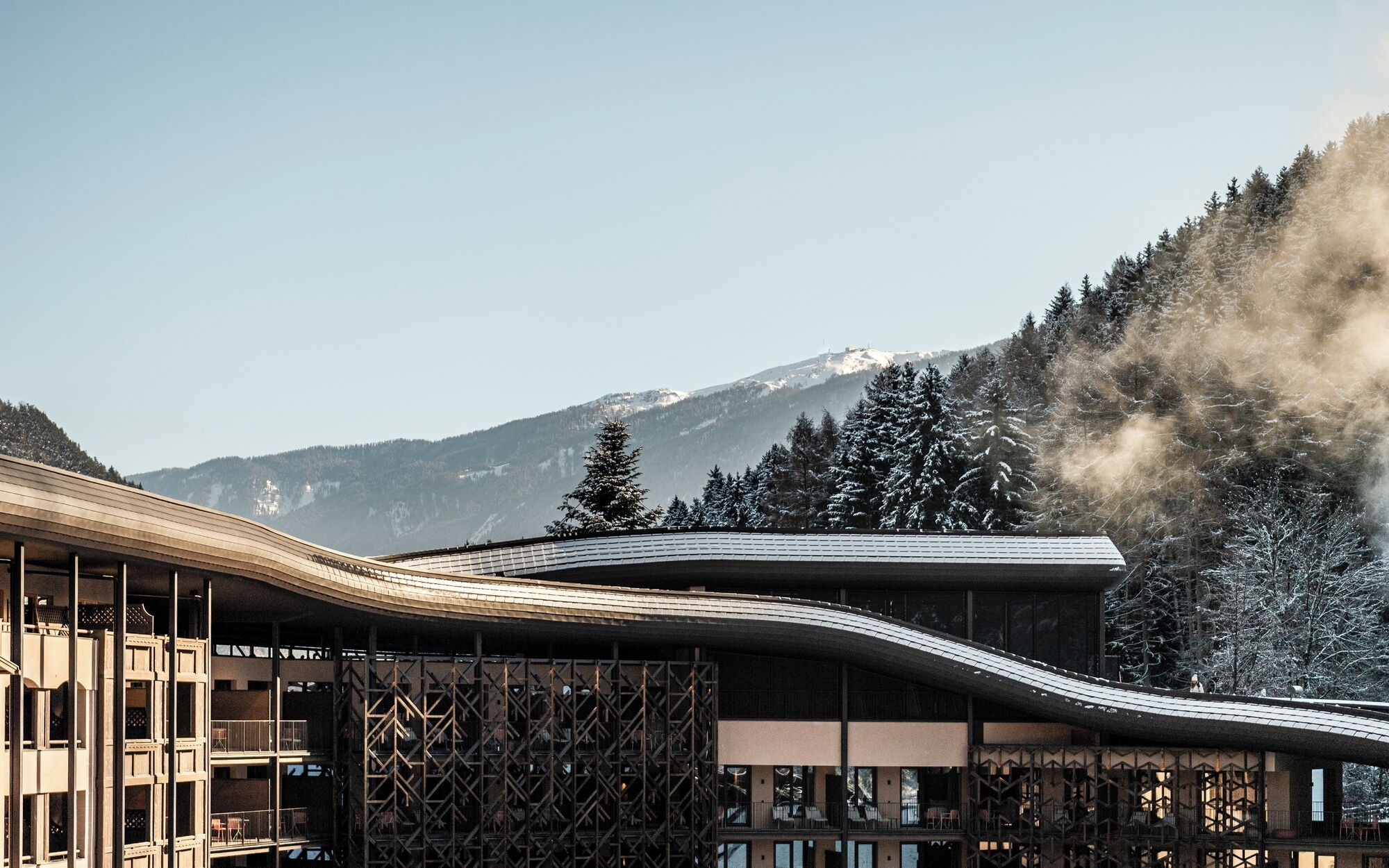 Ein modernes Gebäude mit gewölbter Dachkonstruktion, umgeben von schneebedeckten Bäumen und Bergen im Hintergrund.