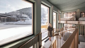 A modern interior with large windows overlooking a snowy landscape, a staircase with wooden railings, and a view of a building with a steam vent. - Falkensteiner Family Resort Lido  (Chienes)
