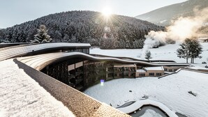 A modern building with a curved roof, surrounded by snow-covered landscape and a forested hill. - Falkensteiner Family Resort Lido  (Chienes)