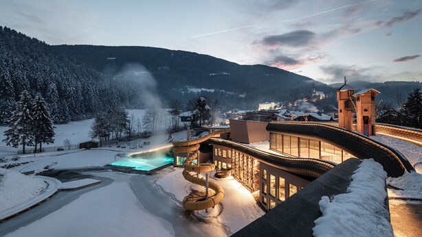 A snow-covered landscape with a modern building featuring a pool, a ski lift, and a church in the background. - Falkensteiner Family Resort Lido  (Chienes)