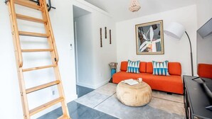 Un salon moderne avec une échelle en bois, un pouf rond, un canapé rouge agrémenté de coussins à motifs, un tableau abstrait encadré et une lampe sur pied.