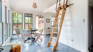 Une pièce spacieuse avec de grandes fenêtres, une échelle en bois et un coin repas comprenant des chaises et une table.