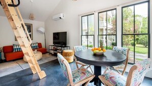 Un salon moderne avec une table ronde, des chaises colorées, une échelle en bois, un canapé et de grandes fenêtres donnant sur l’extérieur.