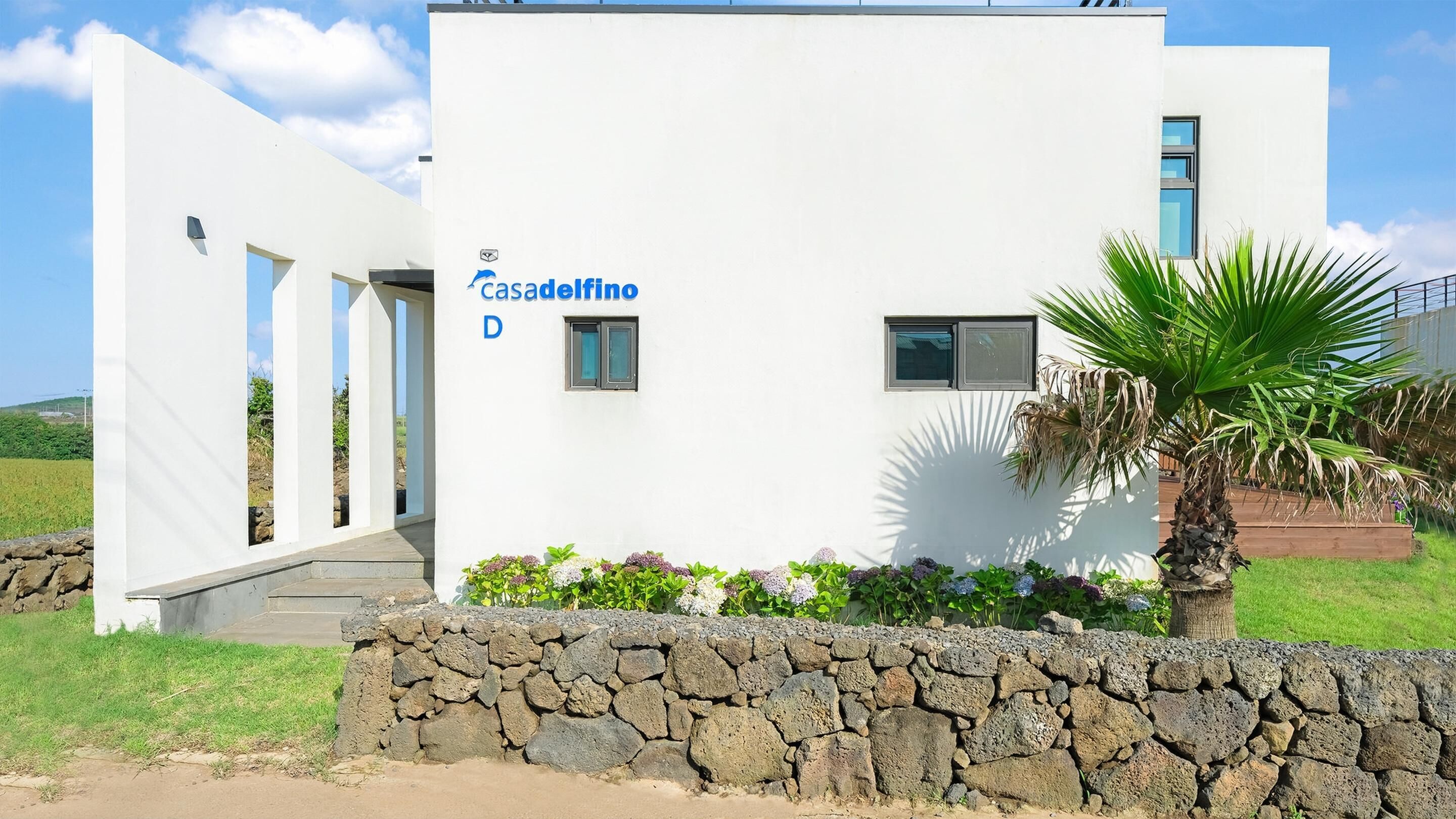 A modern building with a white facade, a stone wall in front, and a palm tree.