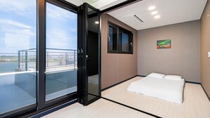 Et moderne hotelværelse med en stor skydedør til en udendørs balkon, en seng med hvide sengetæpper, et lille vindue og et billede på væggen.