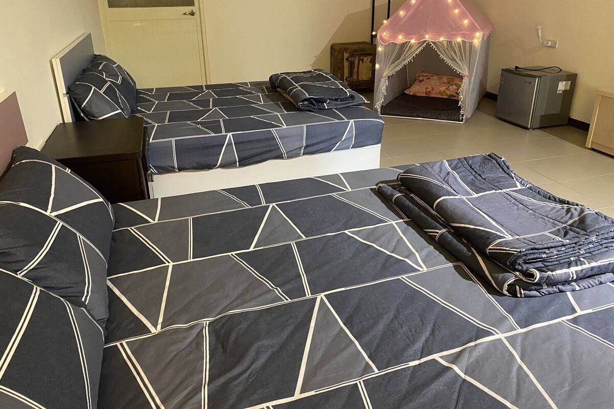 Habitación con dos camas, cada una con cubrecama de estampado geométrico, una cama tipo dosel pequeña con luces, un refrigerador y una maleta.