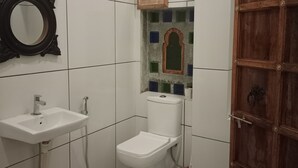 A bathroom with a white sink, toilet, and shower. There is a wooden door and a mirror on the wall. - Lassi Guest House (Udaipur)