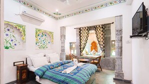 A hotel room with a bed, a small table with chairs, a TV, and decorative wall panels. - Lassi Guest House (Udaipur)