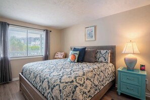 Un dormitorio con cama, mesita de noche, lámpara, un cuadro enmarcado y una ventana con cortinas.