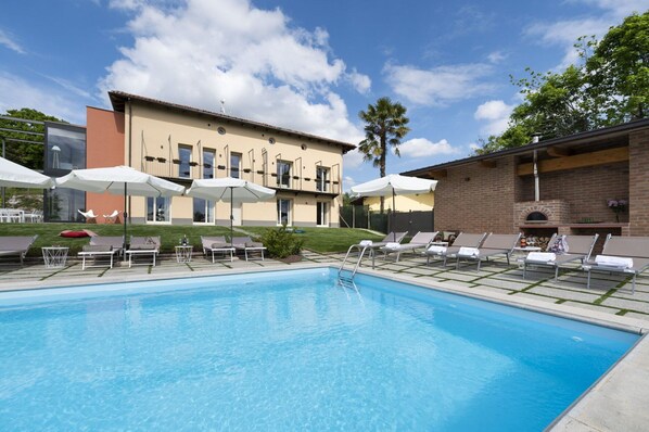 A swimming pool with lounge chairs and umbrellas, a brick building with a barbecue area, and a modern building with large windows. - Private Villa with A/C, WIFI, private pool, TV, patio, pets allowed, panoramic view, close to Alba (San Grato)
