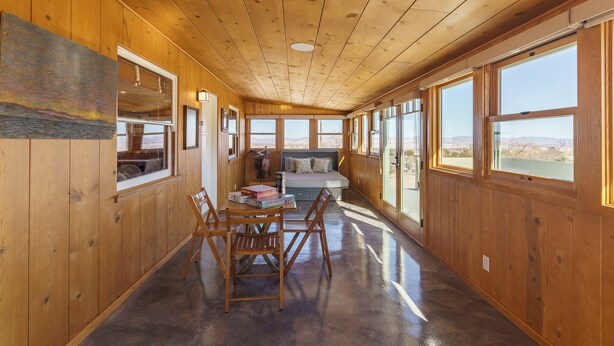 A wooden cabin interior with a dining area, a bed, and large windows offering a view of the landscape.