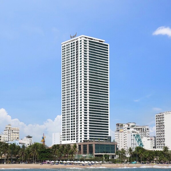 Ein hohes, modernes Hotel mit einer rasterartigen Fassade, nahe einem Strand mit Palmen und anderen Gebäuden im Hintergrund.